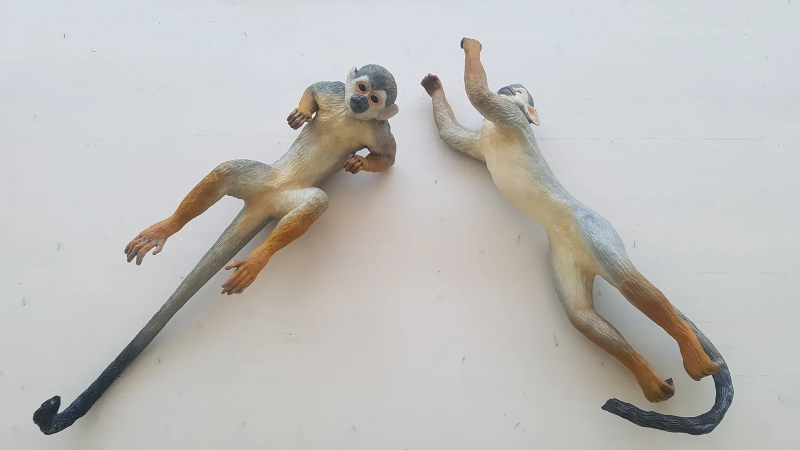 Two epoxy-resin Saimiri sculptures lie face-up on a white surface, showing painted faces, modeled fur, open limbs and extended tails.