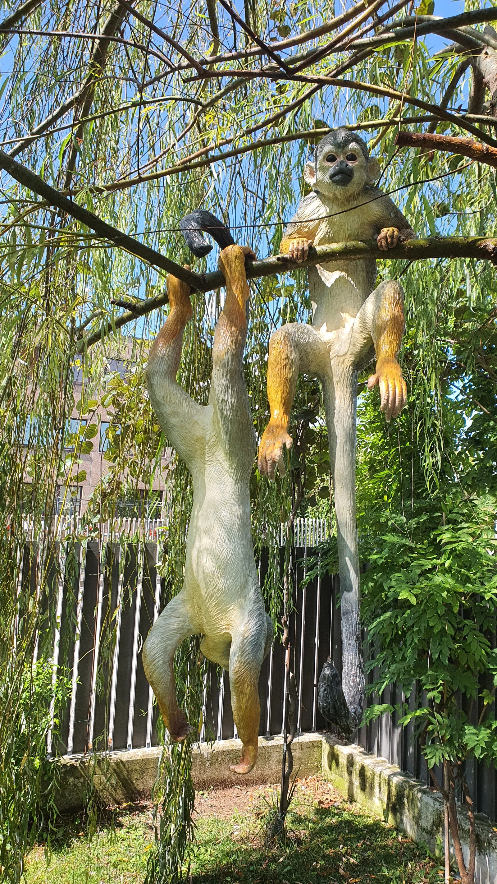 Two painted Saimiri sculptures grip branches among green leaves, with one facing the camera and both extended tails visible.